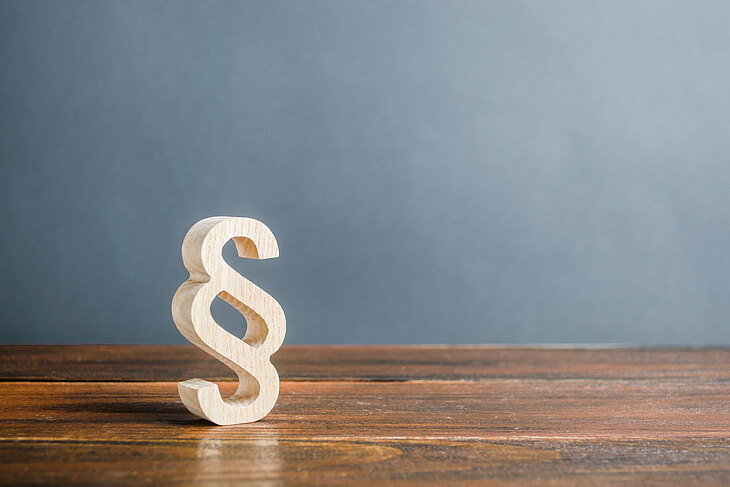 Ein hölzernes Paragraphenzeichen steht auf einem Holztisch vor einer grauen Wand