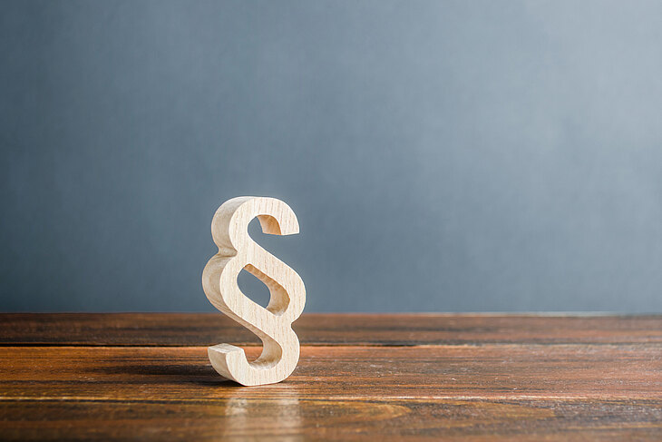Ein hölzernes Paragraphenzeichen steht auf einem Holztisch vor einer grauen Wand