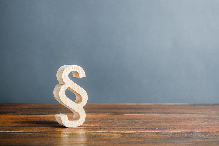 Ein hölzernes Paragraphenzeichen steht auf einem Holztisch vor einer grauen Wand